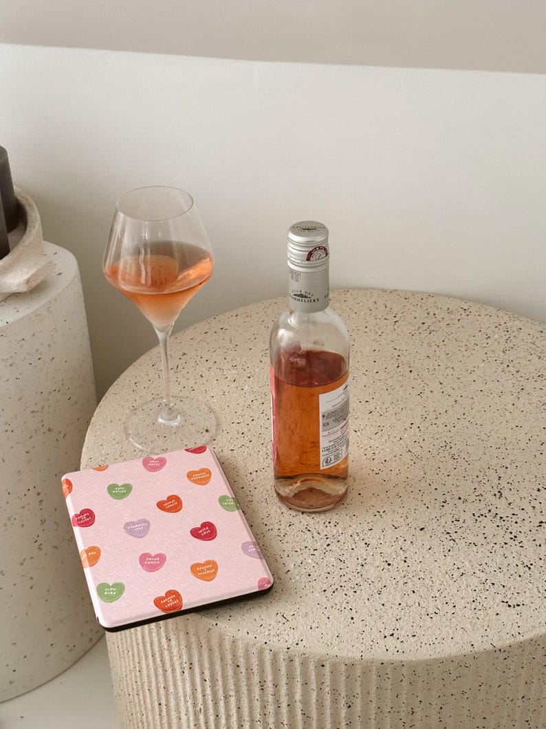 Puede incluir: Un lector electr&oacute;nico rosa con un patr&oacute;n de corazones coloridos, una copa de vino rosado y una botella de vino rosado sobre una superficie beige moteada.