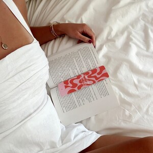 May include: A pink and red bookmark with the text "Sound Girls Read Space" and a wavy abstract design. The bookmark is placed in a book.