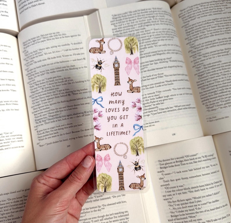 May include: A pink and blue bookmark with a watercolor design featuring a deer, a bee, a clock tower, and a bow. The bookmark has the text "How many loves do you get in a lifetime?" printed on it.