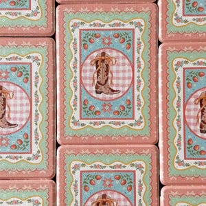 May include: A collection of rectangular decorative tins with a Western theme. Each tin features a cowboy boot image within a circle, surrounded by floral and strawberry motifs, set against a pink and blue backdrop.