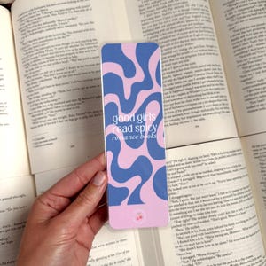 May include: A pink and blue bookmark with a wavy abstract design. The bookmark has the text "good girls read spicy romance books" in white.