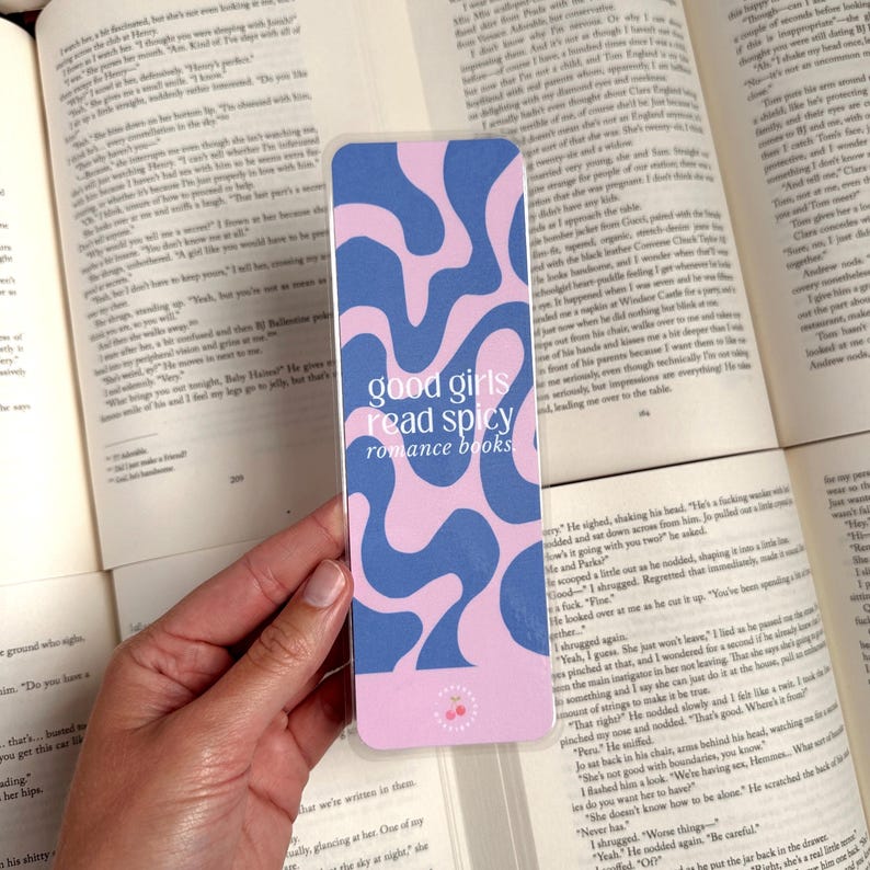 May include: A pink and blue bookmark with a wavy abstract design. The bookmark has the text "good girls read spicy romance books" in white.