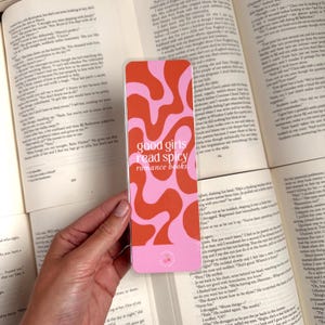 May include: A pink and orange bookmark with a wavy abstract design. The bookmark has the text "good girls read spicy romance books" printed on it.