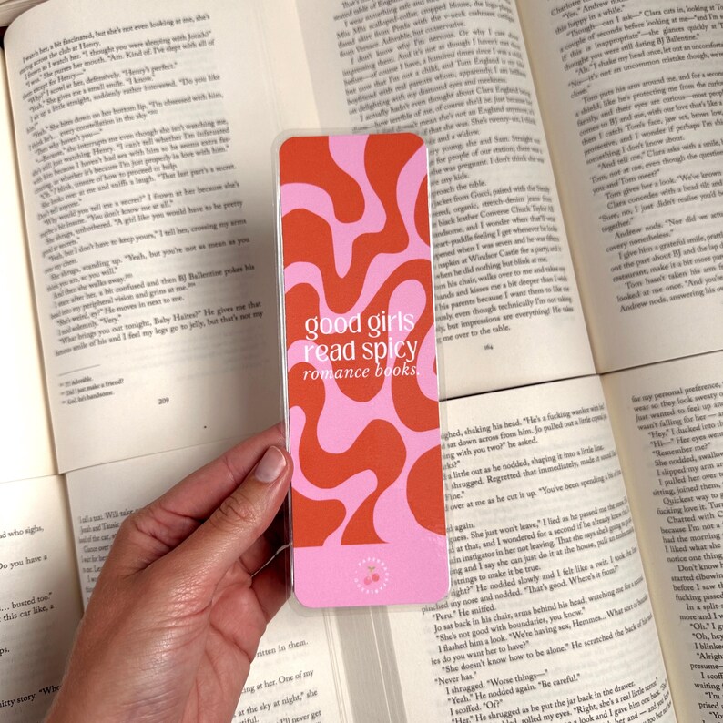 May include: A pink and orange bookmark with a wavy abstract design. The bookmark has the text "good girls read spicy romance books" printed on it.