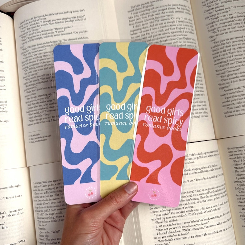 May include: Three colorful bookmarks with a pink, blue, and orange abstract design. The bookmarks have the text "good girls read spicy romance books" on them.