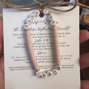 May include: A white beaded bracelet with gold lettering spelling "JOHN 1:16" and "GRACE". The bracelet is on a white card with text about the meaning of the scripture verses.