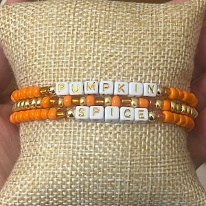 May include: Three beaded bracelets with the words "PUMPKIN SPICE" spelled out in white letter beads. The bracelets are orange and gold, and are displayed on a small, tan burlap pillow.