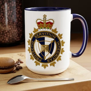 May include: White ceramic coffee mug with a blue rim and handle. The mug features a gold and blue crest with the words "SERVITIUM", "PROTECTIO", "INTEGRITAS", and "VERITAS" around the crest. A crown is above the crest.