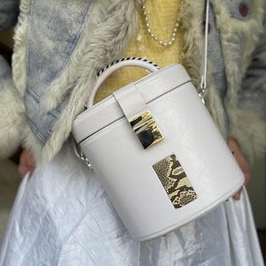 Könnte beinhalten: Eine weiße zylindrische Handtasche mit einem oberen Griff und einem Schulterriemen. Die Tasche hat einen goldfarbenen Verschluss und einen rechteckigen Akzent mit Schlangenmuster. Der Hintergrund zeigt eine hellblaue Jacke mit Fellbesatz und einen weißen Rock.