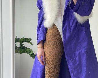 Op de afbeelding: Een lange paarse jas met een witte bontkraag en manchetten. De jas wordt gedragen over een wit bodysuit en bruine luipaardprint panty's.