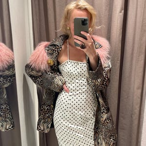 Pode incluir: Um vestido midi branco com bolinhas pretas e alças finas usado sob um casaco de manga comprida com um patchwork de estampas de leopardo e zebra, finalizado com uma gola de pele sintética rosa e grandes botões dourados.