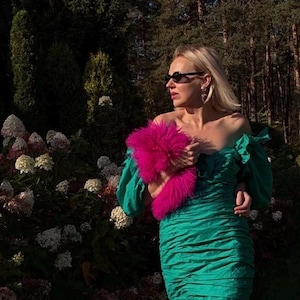 Könnte beinhalten: Ein schulterfreies smaragdgrünes Kleid mit gerafftem Oberteil, Puffärmeln und einem gerüschten Saum, kombiniert mit einem leuchtend pinkfarbenen Kunstfell-Accessoire. Das Kleidungsstück zeichnet sich durch eine strukturierte Stoffoberfläche aus.