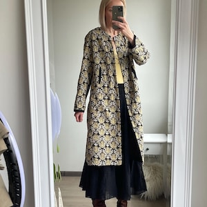 Pode incluir: Um casaco comprido e aberto na frente, com um padrão de damasco preto e dourado. O casaco é usado sobre um top amarelo e uma saia preta. Botas de couro castanho completam o conjunto. O design do casaco apresenta um motivo floral repetido.