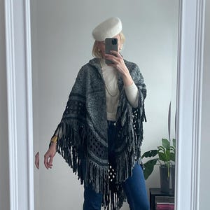 Puede incluir: Un poncho gris con capucha y flecos y una boina blanca se reflejan en un espejo. El poncho tiene un patrón texturizado y flecos grises oscuros en la parte inferior. La boina es de color crema claro.