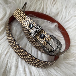 Peut inclure: Une ceinture à motif peau de serpent avec un dos en cuir marron. La ceinture est ornée d'une boucle argentée et de deux rivets décoratifs sur la boucle. La ceinture est enroulée sur une surface blanche et duveteuse.