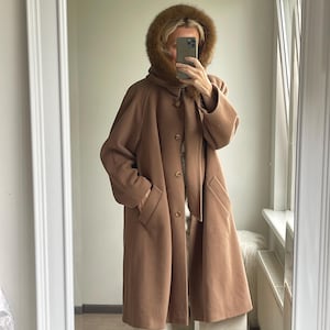 May include: A camel-colored wool coat featuring a hood with a brown faux fur trim, long sleeves, and a front button closure. The coat includes two side pockets and falls to the mid-thigh, worn over light-colored trousers.