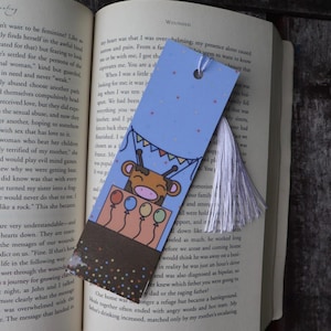 May include: A whimsical bookmark featuring a giraffe in a hot air balloon, holding colorful balloons. The bookmark has a blue background with a tassel and is placed in an open book.