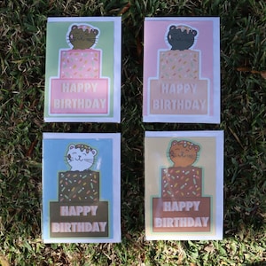 May include: Four rectangular greeting cards featuring illustrations of cats sitting atop tiered birthday cakes with colorful sprinkles. Each card displays the text HAPPY BIRTHDAY in white capital letters. The cards have green, pink, blue, and tan backgrounds.
