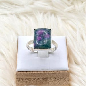 Op de afbeelding: Een zilveren ring met een rechthoekige edelsteen met een mix van groene en paarse tinten. De ring wordt gepresenteerd op een wit oppervlak, tegen een pluizige, witte achtergrond. De edelsteen is het middelpunt en toont zijn unieke kleur en vorm.