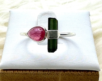 May include: A silver ring with a pink teardrop-shaped tourmaline and a green rectangular tourmaline. The ring is displayed on a white surface with a wooden base.