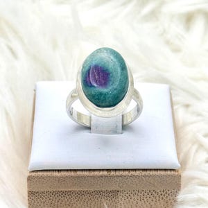 Könnte beinhalten: Ein silberner Ring mit einem ovalen, teal-farbenen und lila Edelstein. Der Ring wird auf einer weißen Oberfläche vor einem flauschigen, weißen Hintergrund präsentiert.