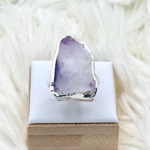 Può includere: Anello argentato con una grande ametista di forma irregolare. La pietra ha una tonalità viola chiaro con aree bianche e traslucide. L'anello è esposto su una superficie bianca, su uno sfondo bianco e soffice.