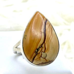 May include: A silver ring with a large, teardrop-shaped stone. The stone displays a warm, earthy colour palette with shades of tan, brown, and cream, and a unique, natural pattern. The ring's band has a simple, split design.