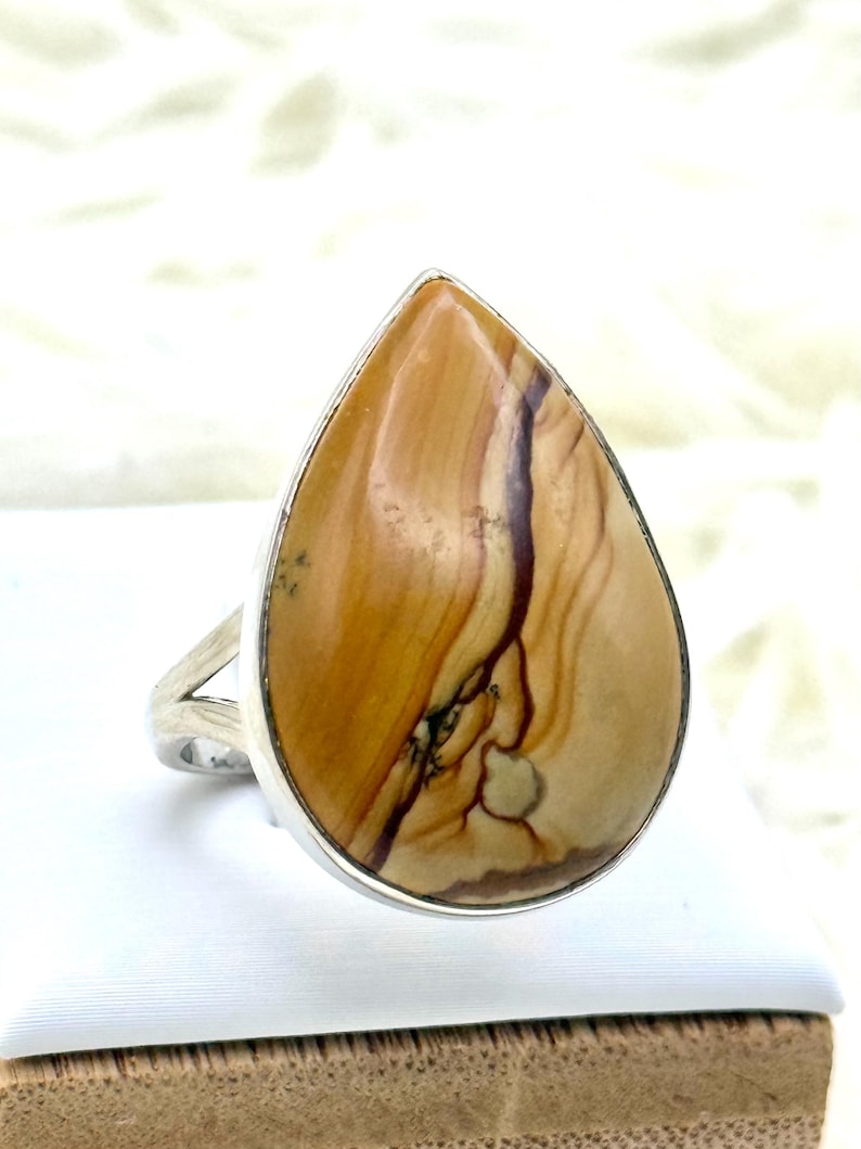 May include: A silver ring with a large, teardrop-shaped stone. The stone displays a warm, earthy colour palette with shades of tan, brown, and cream, and a unique, natural pattern. The ring's band has a simple, split design.