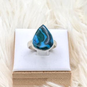 Puede incluir: Un anillo de plata con una piedra en forma de lágrima que presenta un llamativo patrón de tonos azules y verdes. El anillo se exhibe sobre un soporte blanco, colocado sobre una superficie blanca y esponjosa. El color y la forma únicos de la piedra lo convierten en una joya destacada.