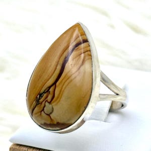 May include: A silver ring with a large, teardrop-shaped stone. The stone has a polished finish and displays a pattern of tan, brown, and black hues. The ring's band has a simple, split design.