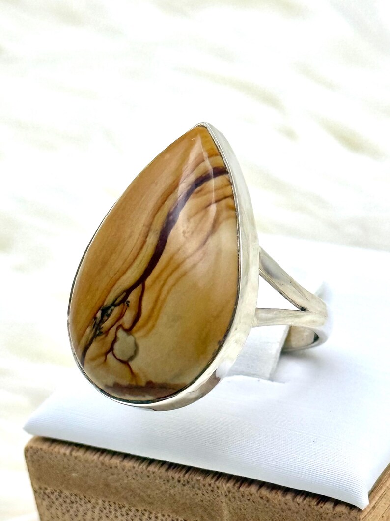 May include: A silver ring with a large, teardrop-shaped stone. The stone has a polished finish and displays a pattern of tan, brown, and black hues. The ring's band has a simple, split design.
