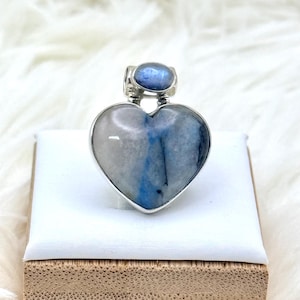 May include: A heart-shaped pendant with a silver-colored frame. The pendant features a blue and white stone with a smaller blue stone at the top. The pendant is displayed on a white surface.