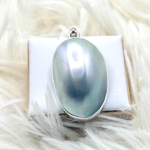 May include: A large, oval-shaped, iridescent gray pearl pendant set in a polished silver frame. The pearl displays a smooth, lustrous surface with subtle blue and green undertones. A small silver bail is attached to the top of the metal setting.