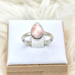 May include: A silver ring featuring a pear-shaped, light pink gemstone. The ring is set on a white surface and presented on a wooden base. The gemstone has a soft, marbled appearance.