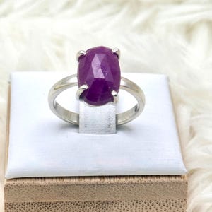 Op de afbeelding: Een zilveren ring met een grote, facetgeslepen, ovale robijn. De diepmagenta steen wordt vastgehouden door vier klauwen. De ring wordt gepresenteerd op een wit oppervlak, tegen een zachte, witte achtergrond.