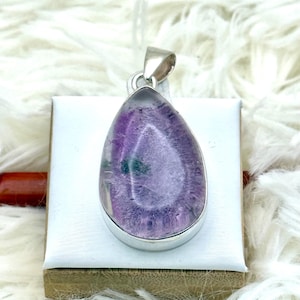 May include: A teardrop-shaped pendant with a silver-colored bezel. The pendant features a translucent purple stone with a lighter center and some green inclusions. The pendant is displayed on a white surface.