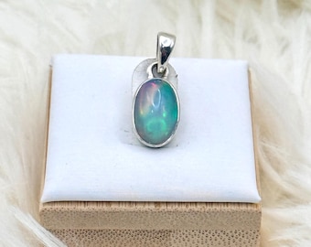 May include: A silver pendant with an oval opal gemstone. The opal displays a play of color with hues of blue, green, and purple. The pendant is displayed on a white surface atop a wooden box, set against a fluffy white background.