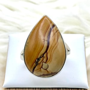 May include: A teardrop-shaped ring with a polished, patterned stone in tan and brown, set in a silver band. The stone has dark brown veining and a unique natural pattern. The ring is displayed on a white surface.