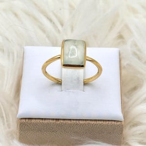 May include: A gold ring featuring a rectangular, pale green, translucent stone set in a smooth metal bezel. The band is thin and circular, crafted from a polished gold-toned metal that contrasts with the soft, muted green hue of the central gemstone.