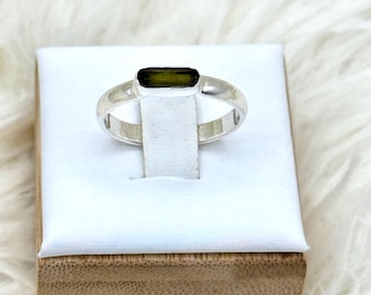 May include: A silver ring featuring an olive-green rectangular gemstone. The ring is presented on a white stand with a wooden base, set against a fluffy white backdrop. The gemstone is the central feature of the ring.