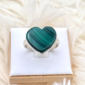 Puede incluir: Anillo de plata con una piedra de malaquita en forma de corazón. La piedra es de color verde intenso con patrones arremolinados. El anillo se muestra sobre una superficie blanca.