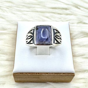 May include: A silver ring with a rectangular, light purple stone. The ring has a decorative design on each side of the stone. The ring is displayed on a white stand with a brown base.