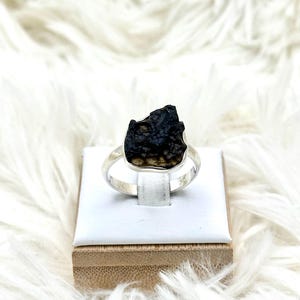 May include: A ring featuring a raw, dark brown and black textured stone set in a polished silver band. The stone has an irregular, jagged shape with a pitted surface, securely held by a smooth metal bezel setting that encircles the base of the gem.