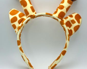 Op de afbeelding: Een grillige giraf-hoofdband met crèmekleurige stof en bruine vlekken. De hoofdband heeft twee rechtopstaande, cilindrische vormen met bruine pompons en twee oren. Dit kostuumaccessoire is perfect voor themafeesten of verkleedpartijen.