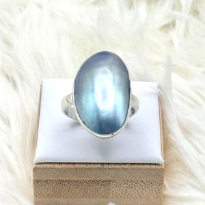 May include: A silver ring featuring a large, oval-shaped, iridescent blue gemstone. The gemstone has a pearlescent sheen and is set in a simple silver band. The ring is displayed on a white surface atop a wooden box.
