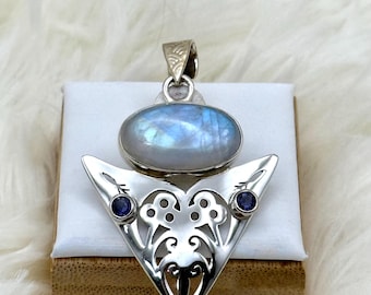 Op de afbeelding: Zilveren hanger met een ovale, iriserende maansteen en twee kleine, ronde blauwe edelstenen. De hanger heeft een decoratief, opengewerkt ontwerp in een driehoekige vorm.