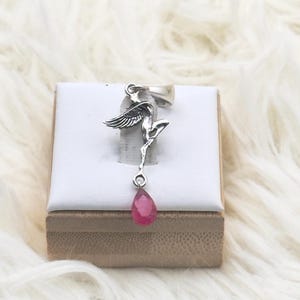 May include: A silver pendant featuring a stylized winged figure in a dynamic pose, suspended from a bail. A faceted, teardrop-shaped pink gemstone hangs from the base of the figure. The pendant is displayed on a small, square white stand.
