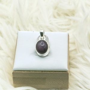 May include: A silver pendant featuring a smooth, oval-shaped dark purple cabochon gemstone. The stone is set in a polished silver bezel with a simple silver bail at the top for attachment to a chain, showcasing a deep, solid color.