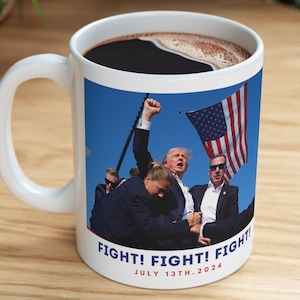 May include: White ceramic mug with a photo of a man in a suit raising his fist in the air. The photo is against a blue sky with a US flag in the background. The text "FIGHT! FIGHT! FIGHT! JULY 13TH, 2024" is printed on the mug.