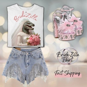 Puede incluir: Camiseta blanca con un gráfico de una novia dinosaurio con velo y sosteniendo un ramo de rosas rosas. El texto "Bridezilla" está impreso en la camiseta. La imagen también muestra un par de tacones de plataforma rosa con detalles de plumas y pedrería, y un par de pantalones cortos de mezclilla con flecos de pedrería.